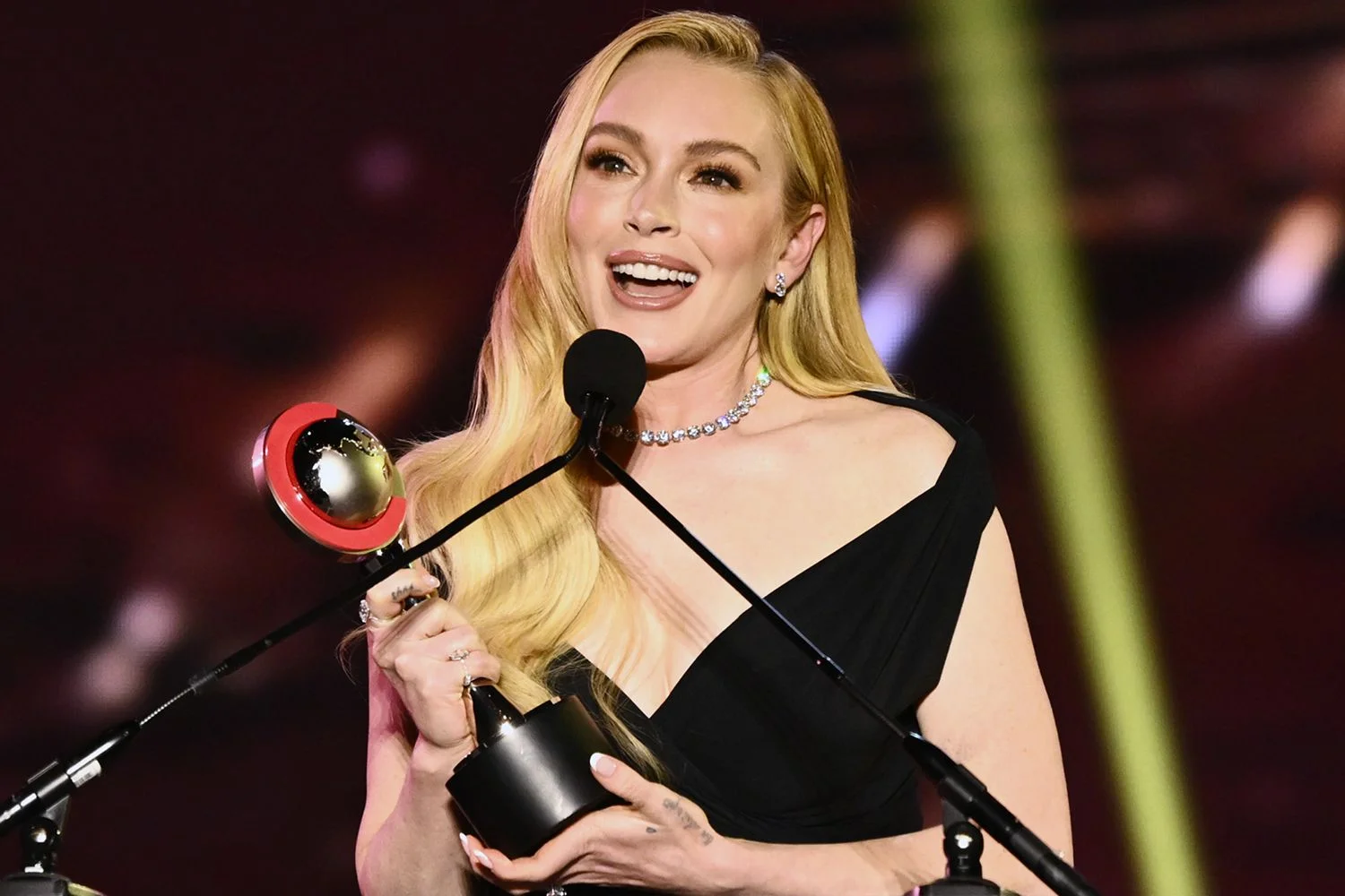 Lindsay Lohan wearing a black off-shoulder Jacquemus gown onstage at CinemaCon 2025 holding her award