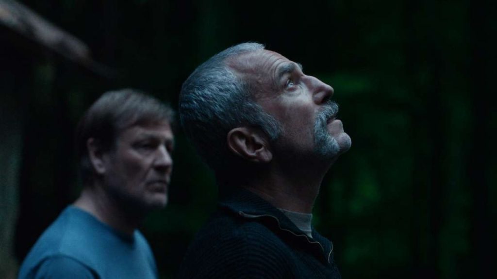 Daniel Day-Lewis standing in a dimly lit forest scene from Anemone, capturing the film’s dramatic tone.