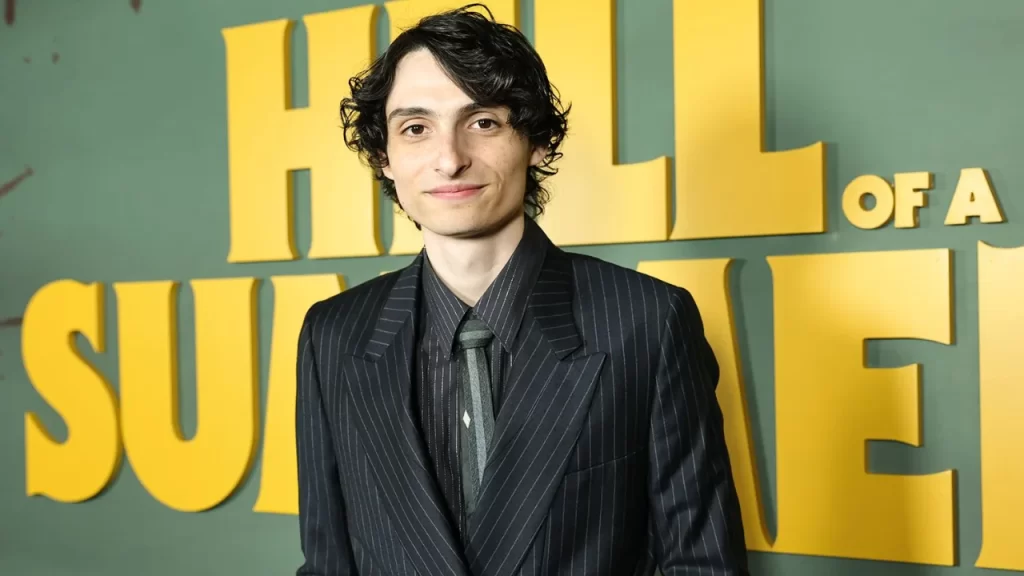 Finn Wolfhard wearing casual camp-style outfit, holding camera equipment on the set of Hell of a Summer, smiling while directing a scene.
