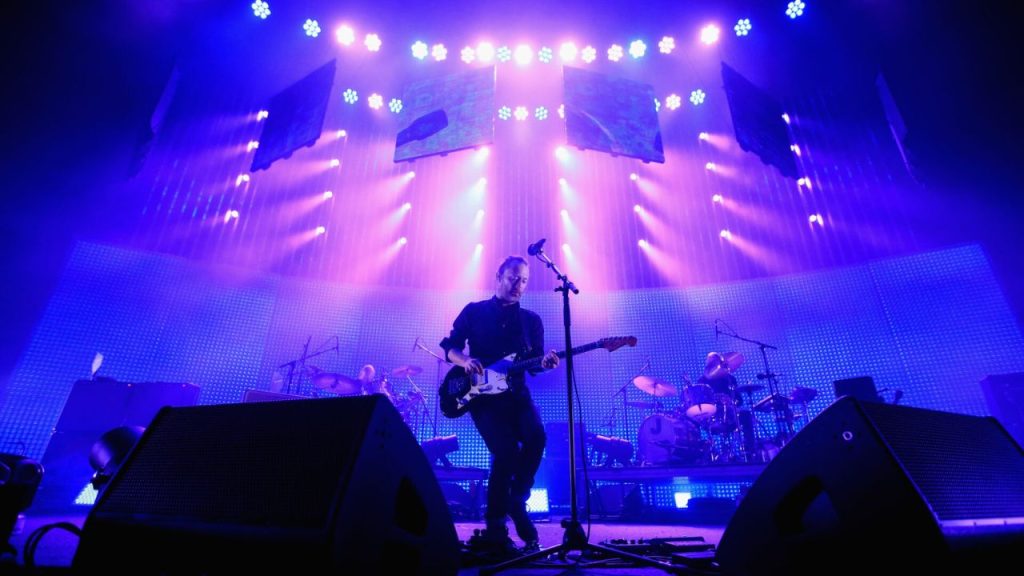 Radiohead performing live on stage, backlit by vibrant purple lighting.