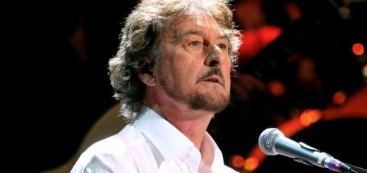 A close-up of Rick Davies singing into a microphone and playing piano during a live concert performance.