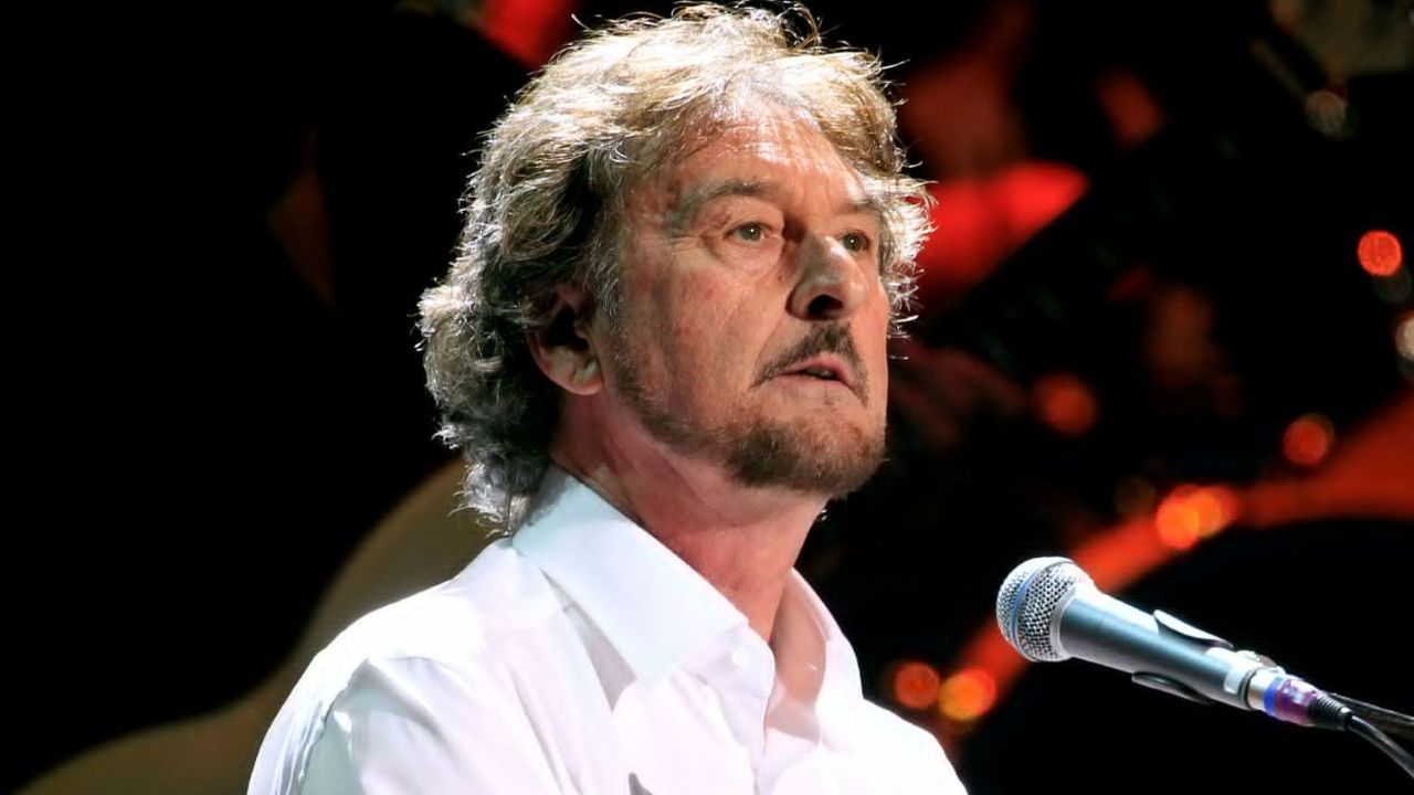 A close-up of Rick Davies singing into a microphone and playing piano during a live concert performance.