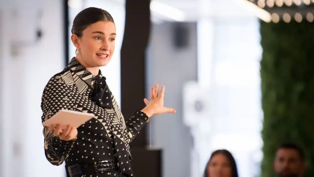 Chloe Malle mid-speech or presentation, wearing a patterned outfit and gesturing—capturing her leadership and public-speaking style.